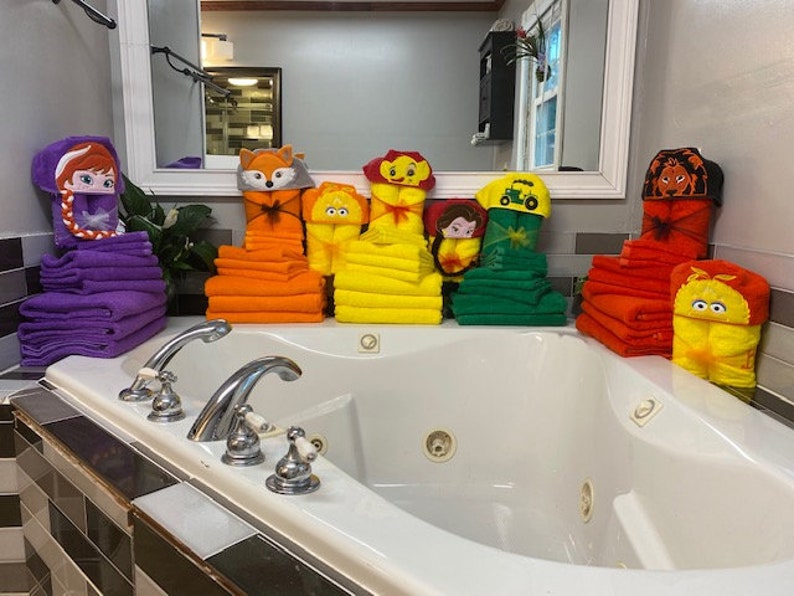 Peut inclure: Une collection de serviettes &agrave; capuche color&eacute;es avec diff&eacute;rents personnages de dessins anim&eacute;s, dont un renard, un lion, une princesse, un tracteur et un oiseau. Les serviettes sont empil&eacute;es dans une salle de bain avec une baignoire blanche.