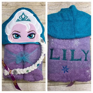 Puede incluir: Toalla con capucha morada con una trenza blanca y una capucha azul. La parte delantera de la toalla tiene una imagen bordada de una ni&ntilde;a con una corona y el nombre "LILY" est&aacute; bordado en la parte posterior.