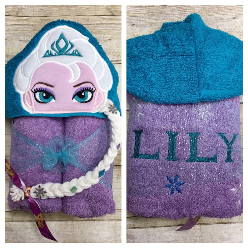 Puede incluir: Toalla con capucha morada con una trenza blanca y una capucha azul. La parte delantera de la toalla tiene una imagen bordada de una ni&ntilde;a con una corona y el nombre "LILY" est&aacute; bordado en la parte posterior.