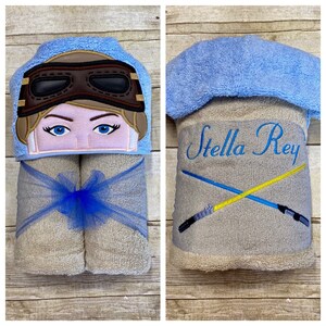 May include: A light blue hooded towel with a girl's face embroidered on the hood. The towel is rolled up and tied with a blue ribbon. The other side of the towel has the name "Stella Rey" and two crossed lightsabers embroidered on it.