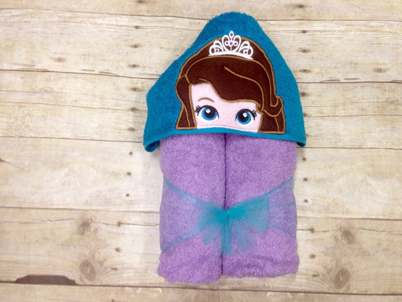 May include: A purple hooded towel with a turquoise hooded top featuring an embroidered princess with a crown. The towel has a turquoise ribbon tied around it.