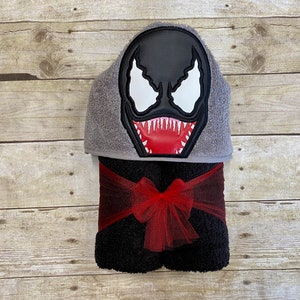 Puede incluir: Una toalla con capucha negra con un personaje de Venom bordado en rojo y blanco en la capucha. La toalla est&aacute; atada con una cinta roja.