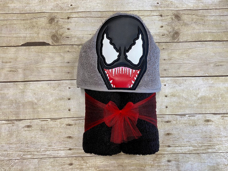 Puede incluir: Una toalla con capucha negra con un personaje de Venom bordado en rojo y blanco en la capucha. La toalla est&aacute; atada con una cinta roja.
