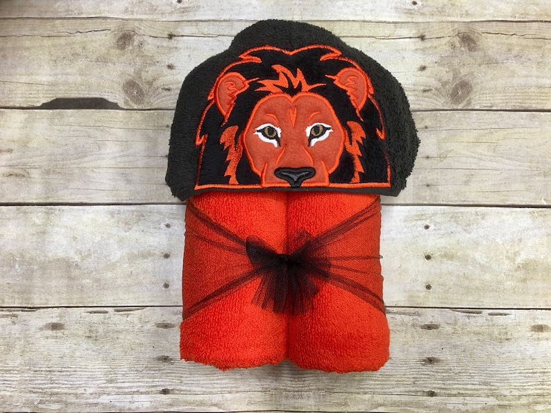 Peut inclure: Serviette &agrave; capuche orange avec un visage de lion noir brod&eacute; sur la capuche. La serviette est nou&eacute;e avec un ruban noir.