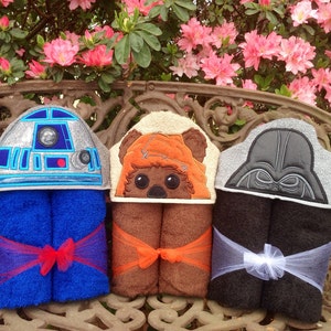 May include: Three hooded towels featuring embroidered Star Wars characters. The towels are folded and tied with ribbon. The towels are displayed on a wooden bench in front of a pink flowering bush.