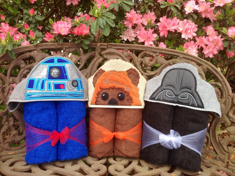 May include: Three hooded towels featuring embroidered Star Wars characters. The towels are folded and tied with ribbon. The towels are displayed on a wooden bench in front of a pink flowering bush.