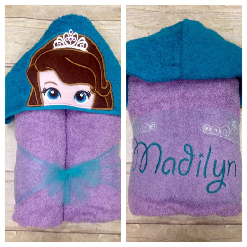 May include: Purple hooded towel with a turquoise trim and a cartoon princess face embroidered on the hood. The towel has a turquoise ribbon tied around it and the name "Madilyn" is embroidered on the back.