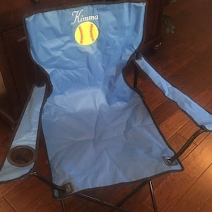 Pu&ograve; includere: Una sedia pieghevole blu con una struttura nera e un portabevande. La sedia ha un design ricamato personalizzato con una palla da softball e il nome "Kimma".