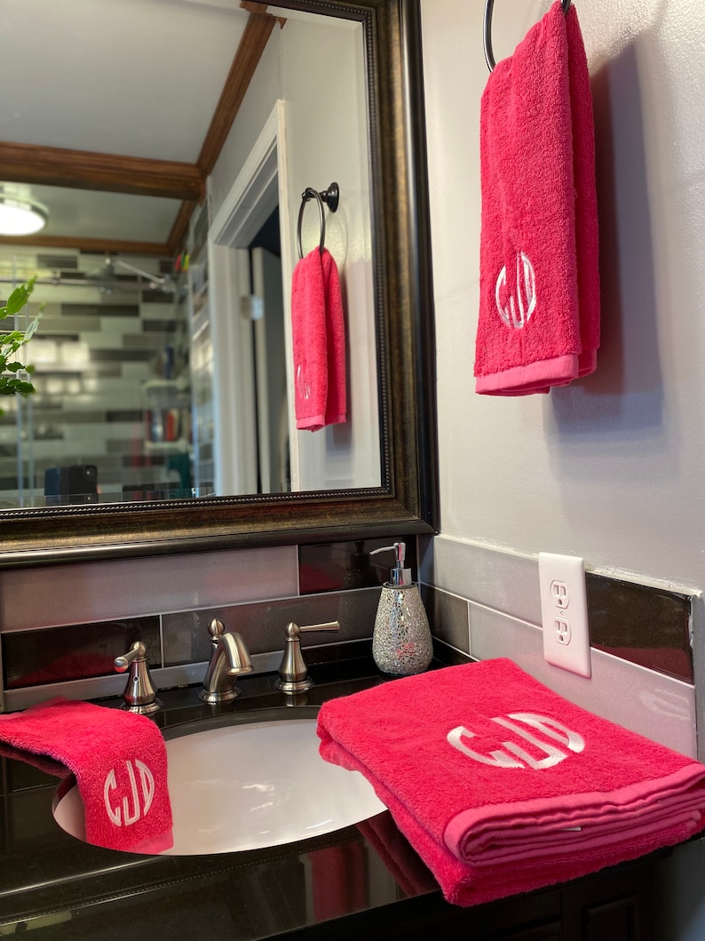 Pu&ograve; includere: Due asciugamani rosa con iniziali ricamate bianche "CJD" appese a un portasciugamani in un bagno. Un terzo asciugamano &egrave; piegato sul bancone del bagno.