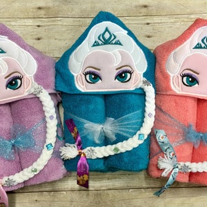 Puede incluir: Tres toallas con capucha con im&aacute;genes bordadas de una princesa con una corona y el pelo trenzado. Las toallas son de color rosa, turquesa y coral. Cada toalla tiene una correa trenzada blanca con pedrer&iacute;a.