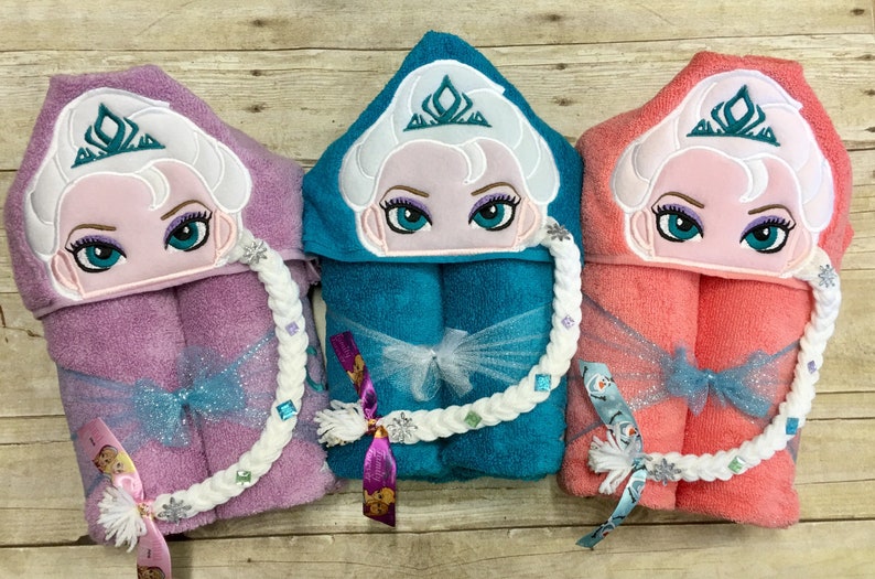 Puede incluir: Tres toallas con capucha con im&aacute;genes bordadas de una princesa con una corona y el pelo trenzado. Las toallas son de color rosa, turquesa y coral. Cada toalla tiene una correa trenzada blanca con pedrer&iacute;a.