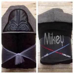 May include: Black hooded towel with a Darth Vader embroidery on the hood and a white tulle bow. The towel is folded and shows the embroidered name "Mikey" and two crossed lightsabers.