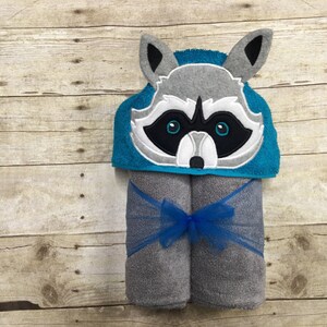 May include: A gray and white raccoon hooded towel with blue trim. The towel is rolled up and tied with a blue ribbon.