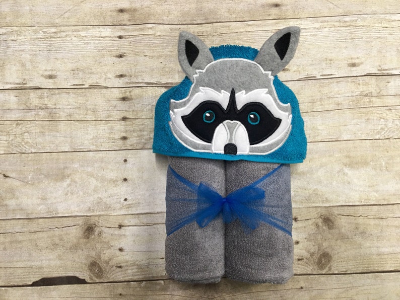May include: A gray and white raccoon hooded towel with blue trim. The towel is rolled up and tied with a blue ribbon.
