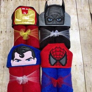 Puede incluir: Cuatro toallas con capucha con dise&ntilde;os de personajes de superh&eacute;roes. Las toallas son rojas, negras, azules y azules con acentos rojos. Cada toalla tiene un personaje diferente: Iron Man, Batman, Superman y Spiderman.