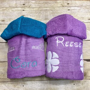 May include: Two purple hooded towels with embroidered names. The towel on the left has a crown and the name "Cora" embroidered in teal. The towel on the right has a white flower and the name "Reese" embroidered in white.