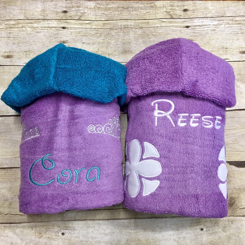 May include: Two purple hooded towels with embroidered names. The towel on the left has a crown and the name "Cora" embroidered in teal. The towel on the right has a white flower and the name "Reese" embroidered in white.