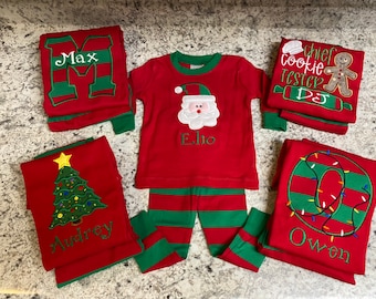 May include: Set of five red and green Christmas pajamas with personalized embroidery. The pajamas feature a Christmas tree, Santa Claus, a gingerbread man, and the letters "M", "O", and "E".