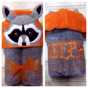 May include: A gray hooded towel with an orange raccoon face on the hood. The towel is tied with an orange ribbon and has the name "JOSEPH" embroidered on the back.