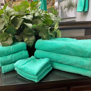 May include: A set of bright teal bath towels, hand towels, and washcloths neatly folded in stacks. The towels feature a soft, textured fabric surface. A large green leafy plant is positioned directly behind the stacks of folded towels.