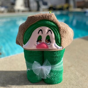May include: A green hooded towel with a brown trim and an embroidered face. The face has brown eyes, a pink nose, and a green hat. The towel is tied with a white ribbon.