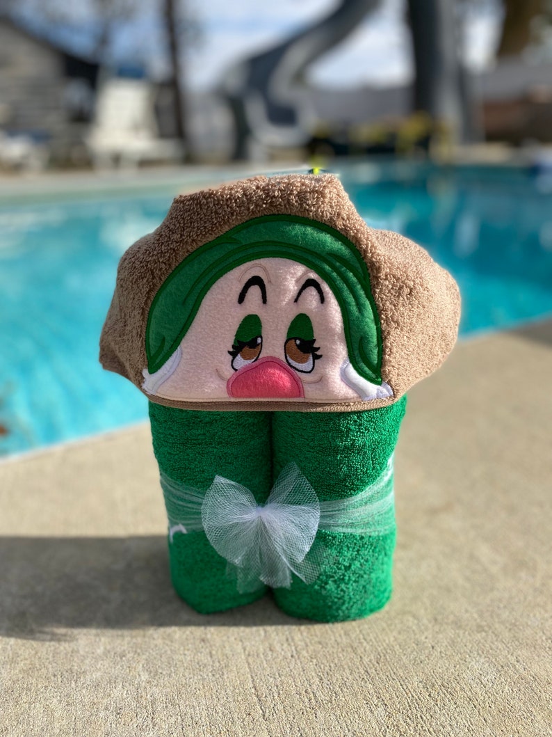 May include: A green hooded towel with a brown trim and an embroidered face. The face has brown eyes, a pink nose, and a green hat. The towel is tied with a white ribbon.