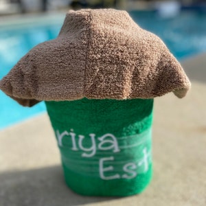 May include: A green towel with the name "Riya Est" embroidered in white. The towel is rolled up and topped with a brown towel.