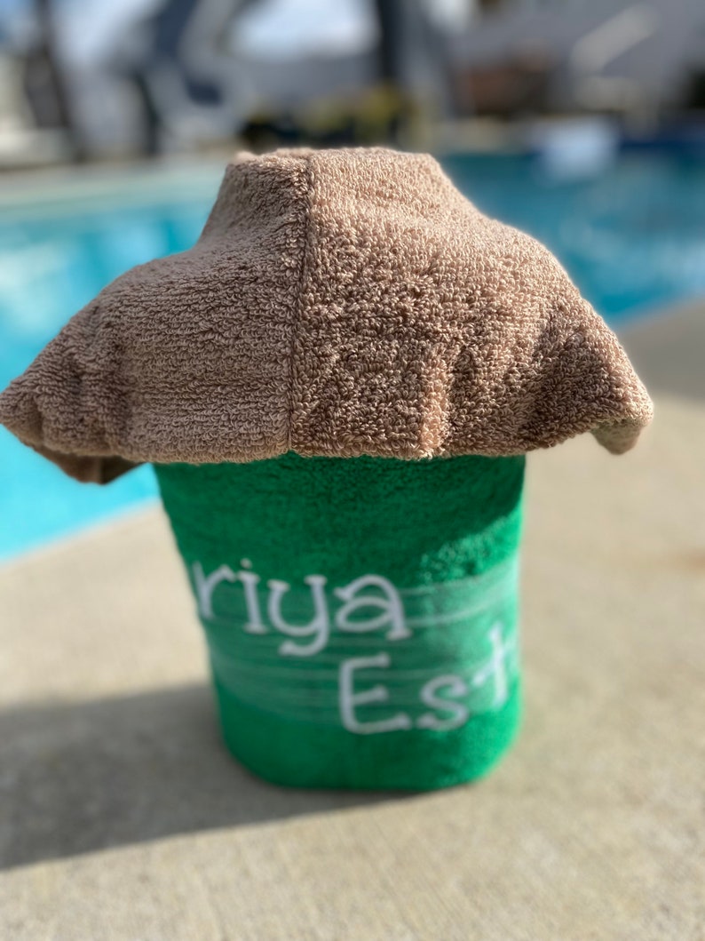 May include: A green towel with the name "Riya Est" embroidered in white. The towel is rolled up and topped with a brown towel.