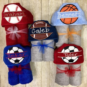 May include: Six hooded towels with different sports themes. Each towel features a different sport ball, including baseball, football, soccer, and basketball. The towels are tied with ribbons and have names embroidered on them, including Graham, Caleb, Tucker, Tyler, and Bennett.