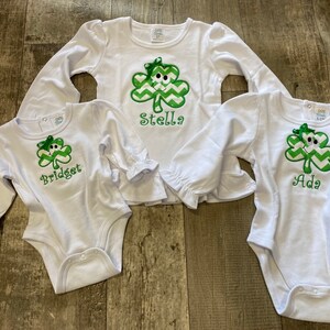 May include: Three white long-sleeved shirts with green shamrock designs and personalized names: Bridget, Stella, and Ada.
