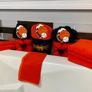Peut inclure: Ensemble de trois serviettes &agrave; capuche noires avec des ballons de sport brod&eacute;s orange et blancs, dont un ballon de basket, un ballon de football et un ballon de baseball. Les serviettes sont pr&eacute;sent&eacute;es avec des serviettes de toilette orange.