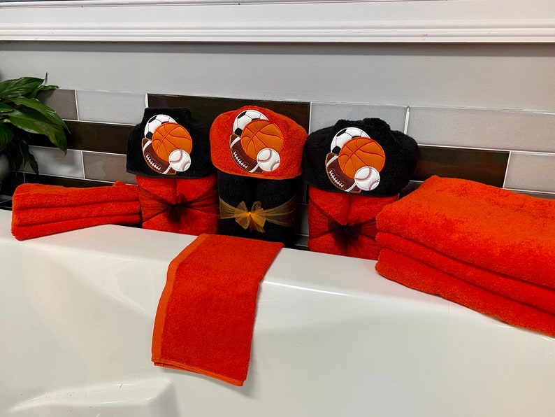 Peut inclure: Ensemble de trois serviettes &agrave; capuche noires avec des ballons de sport brod&eacute;s orange et blancs, dont un ballon de basket, un ballon de football et un ballon de baseball. Les serviettes sont pr&eacute;sent&eacute;es avec des serviettes de toilette orange.