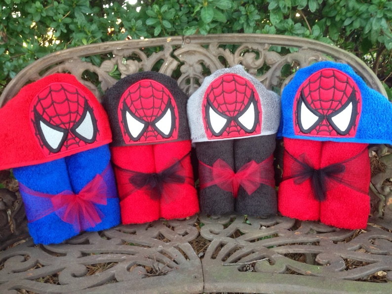 Puede incluir: Cuatro toallas con capucha con un dise&ntilde;o de Spiderman rojo y negro. Las toallas est&aacute;n dobladas y atadas con una cinta negra. Las toallas se muestran en un banco de madera.