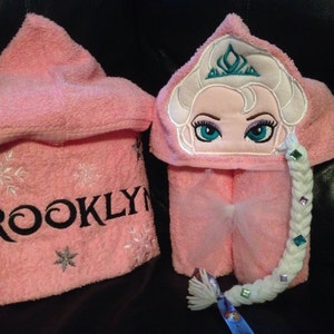 Puede incluir: Toalla con capucha rosa con copos de nieve bordados y el nombre "BROOKLYN". La capucha presenta una imagen bordada de Elsa de Frozen con una trenza y una corona.