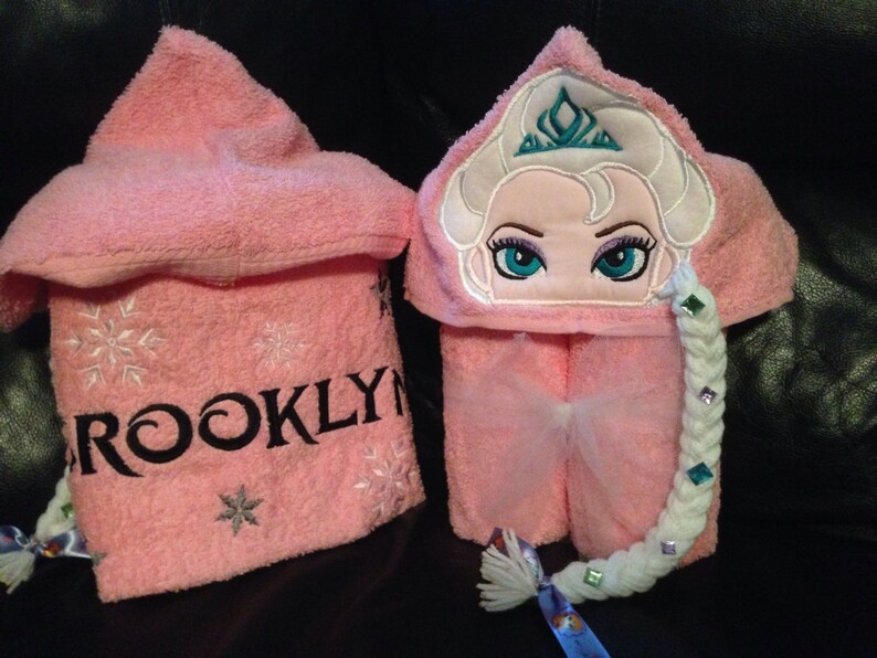 Puede incluir: Toalla con capucha rosa con copos de nieve bordados y el nombre "BROOKLYN". La capucha presenta una imagen bordada de Elsa de Frozen con una trenza y una corona.