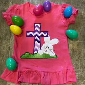 K&ouml;nnte beinhalten: Ein rosa Kleid zum Thema Ostern mit einem Kreuz- und Hasenaufn&auml;her. Das Kleid hat R&uuml;schen&auml;rmel und -saum und ist von bunten Plastik-Ostereiern umgeben. Das Kreuz ist lila und wei&szlig;, und der Hase ist wei&szlig; mit einem l&auml;chelnden Gesicht.