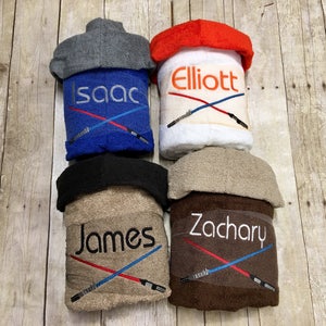 May include: Four hooded towels with personalised names and a crossed lightsaber design. The towels are in blue, white, brown, and beige.