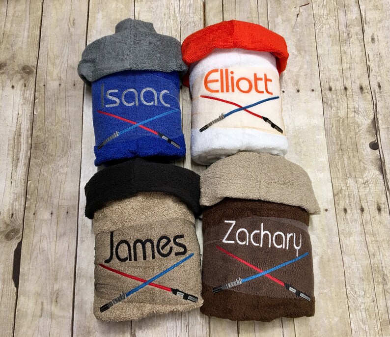 May include: Four hooded towels with personalised names and a crossed lightsaber design. The towels are in blue, white, brown, and beige.