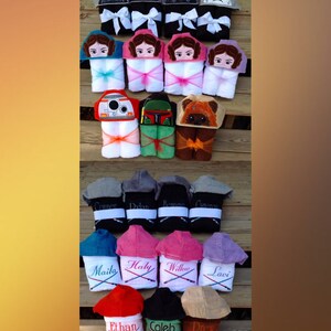 May include: A collection of hooded towels featuring various Star Wars characters embroidered on them. The towels are arranged in rows on a wooden surface. Some towels have names embroidered on them, such as "Connor", "Dylan", "Maile", and "Ethan".