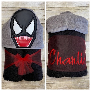 Puede incluir: Una toalla con capucha negra con un lazo rojo y un personaje de Venom bordado en la capucha. La toalla tiene una franja roja y negra y el nombre "Charlie" bordado en la parte inferior.