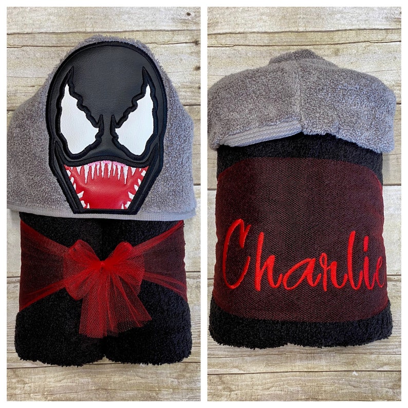 Puede incluir: Una toalla con capucha negra con un lazo rojo y un personaje de Venom bordado en la capucha. La toalla tiene una franja roja y negra y el nombre "Charlie" bordado en la parte inferior.