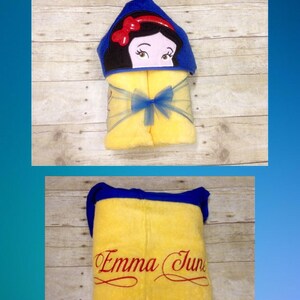 May include: Yellow hooded towel with a blue trim and a Snow White character embroidered on the hood. The towel is folded and tied with a blue ribbon. The towel is personalized with the name "Emma June" in red embroidery.