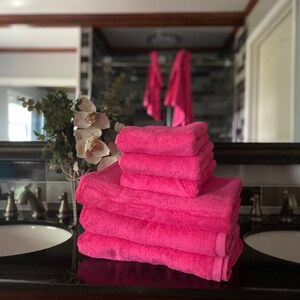 Pu&ograve; includere: Un mucchio di sei asciugamani da bagno rosa piegati ordinatamente su un piano di lavoro marrone scuro in un bagno.