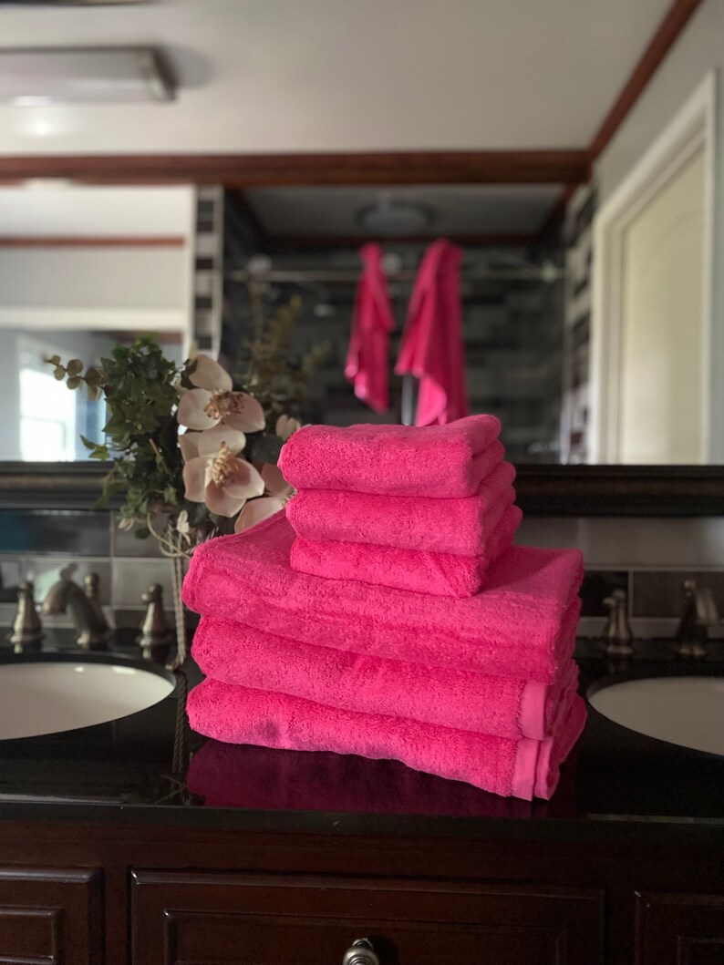 Pu&ograve; includere: Un mucchio di sei asciugamani da bagno rosa piegati ordinatamente su un piano di lavoro marrone scuro in un bagno.