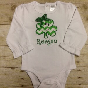 May include: White long-sleeved baby onesie with a green shamrock with a smiling face and the name "Reagan" embroidered on the front.