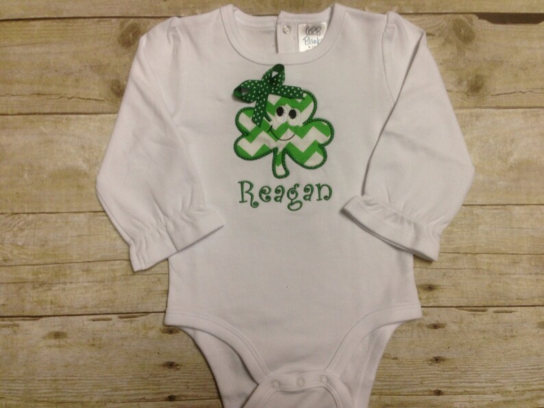 May include: White long-sleeved baby onesie with a green shamrock with a smiling face and the name "Reagan" embroidered on the front.
