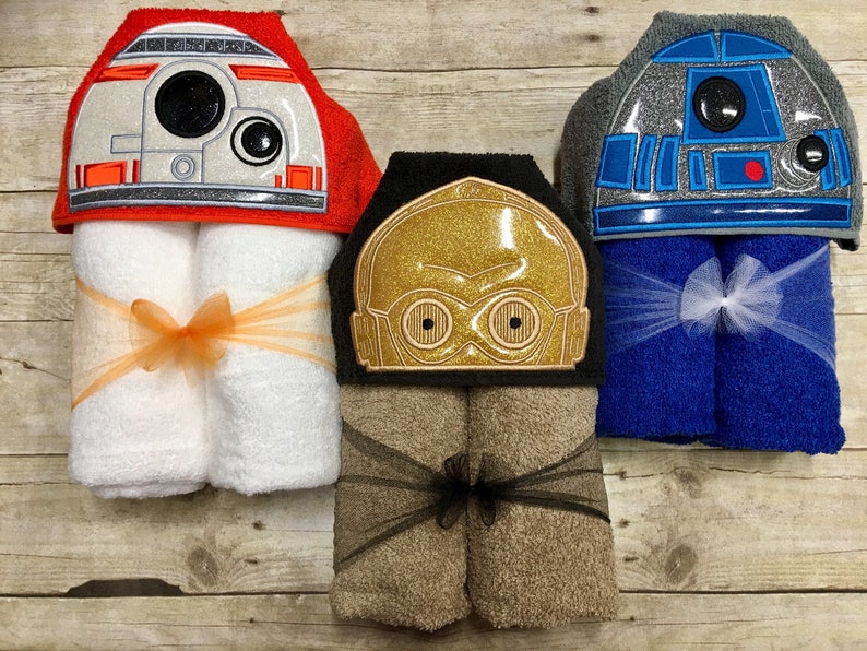 May include: Three hooded towels featuring embroidered Star Wars characters. The towels are rolled up and tied with ribbon. The characters are R2-D2, C-3PO, and BB-8.