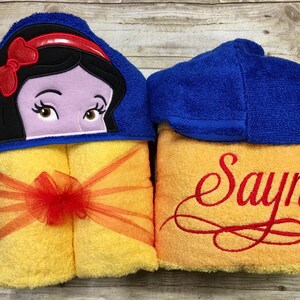 May include: A yellow hooded towel with a blue hood featuring an embroidered image of Snow White. The towel is tied with a red ribbon and has the name "Sayna" embroidered on the bottom.