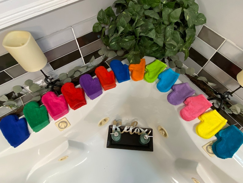 Pu&ograve; includere: Un set di 10 guanti da bagno colorati in un arcobaleno di colori, tra cui blu, verde, rosa, rosso, arancione, giallo, viola e turchese. I guanti sono disposti a semicerchio attorno al bordo di una vasca da bagno bianca. I guanti sono realizzati in un tessuto morbido e testurizzato e sono perfetti per esfoliare e pulire la pelle.