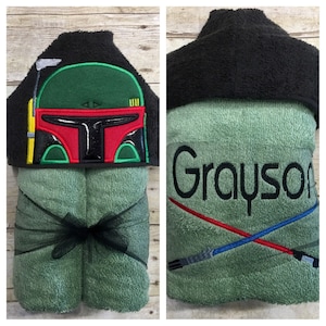 May include: A green hooded towel with a black hood and a green and black embroidered image of a Star Wars character's helmet. The towel is personalized with the name "Grayson" and an embroidered image of two lightsabers.